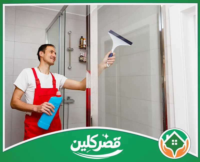 شركة تنظيف الحمامات بالرياض 0550575548 قصر كلين عامل بزي أحمر ينظف زجاج كابينة الاستحمام بالحمام باستخدام مساحة زجاج يدوية ومنظف.