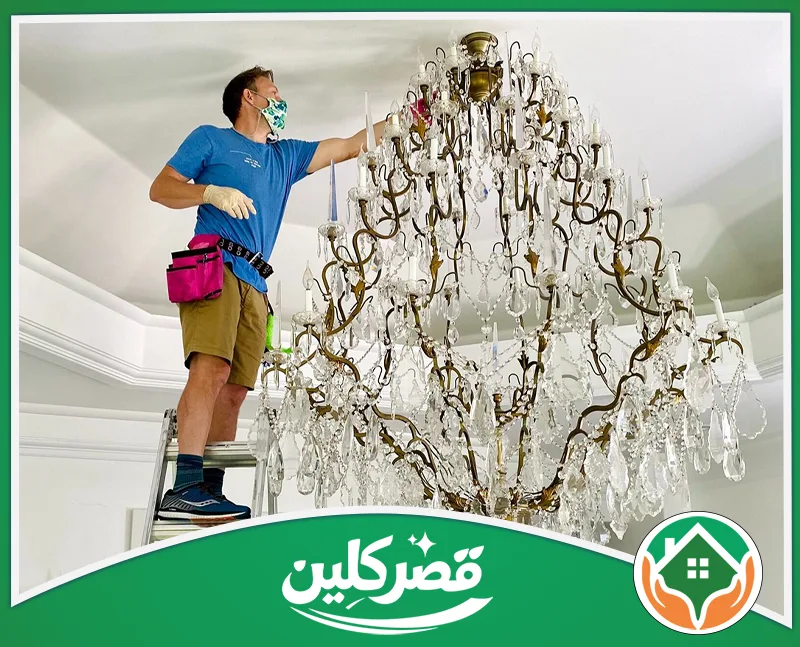 تنظيف النجف المذهب بالرياض 0550575548 قصر كلين صورة لرجل يرتدي كمامة وقفازات، يقف على سلم مرتفع ويقوم بتنظيف نجفة كريستال ضخمة وفاخرة بدقة وعناية.