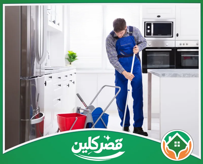 تنظيف خزائن المطابخ بالرياض من الدهون 0550575548 قصر كلين عامل تنظيف يرتدي وزرة زرقاء يمسح أرضية مطبخ أبيض وعصري بعناية، مستعيناً بممسحة يدوية وعربة أدوات التنظيف.