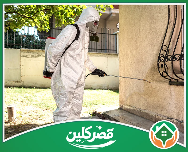 شركة رش مبيدات بالرياض 0550575548 قصر كلين شخص يرتدي بدلة وقاية بيضاء وقناع غاز، يقوم برش المبيدات على جدار مبنى خارجي لمكافحة الآفات.