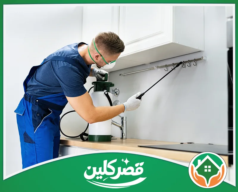 أسعار مكافحة الحشرات بالرياض - قصر كلين عامل يرتدي معدات وقاية أثناء توجيه أداة الرش إلى المنطقة الواقعة أسفل خزائن المطبخ
