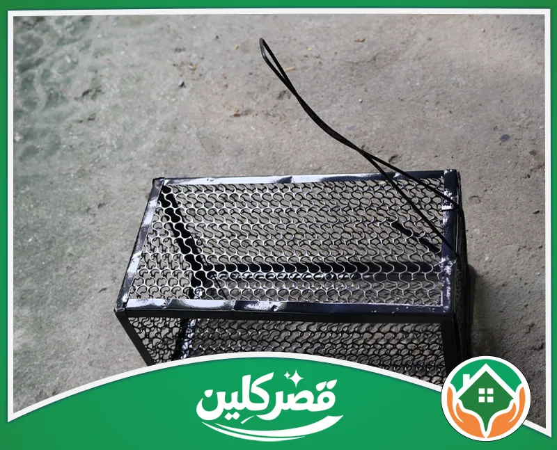 رش الفئران بالرياض 0550575548 قصر كلين مصيدة فئران حديدية سوداء فارغة مصنوعة من الشبك المعدني، موضوعة على أرضية خرسانية رمادية مكشوفة.